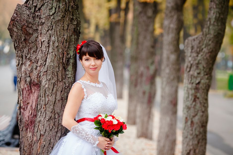 A bride in a white lace wedding dress with delicate floral appliqué details, standing outdoors beside a large tree trunk, holding a vibrant bouquet of red and white roses with green leaves. She wears