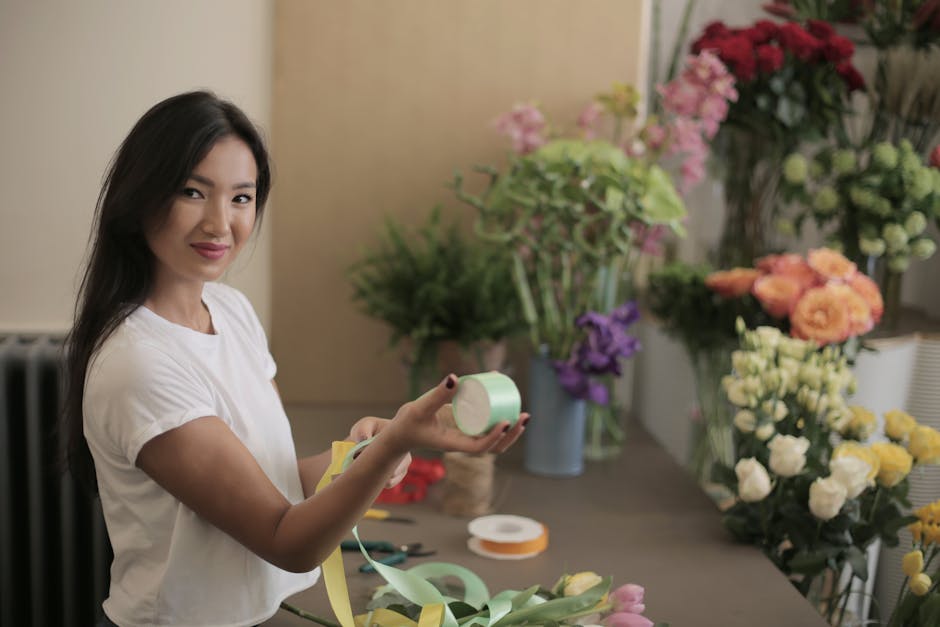 A young woman with long dark hair, wearing a white t-shirt, is smiling while holding a roll of light green floral tape in one hand and a pair of yellow scissors in the other. She is standing in a well