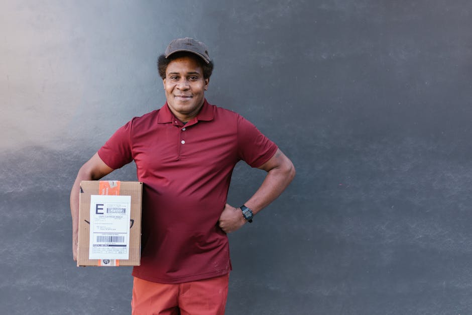 A smiling delivery person with medium brown skin, wearing a maroon polo shirt, matching cap, and smartwatch, stands against a plain, dark grey wall. They are holding a rectangular cardboard box with a