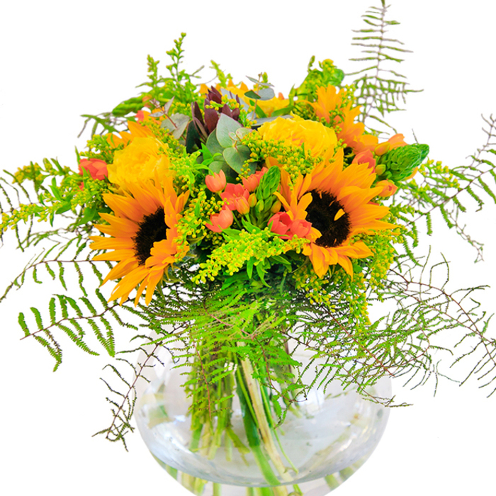 An impression of sunlit exuberance radiates from this opulent bouquet, thoughtfully designed to mirror the sunny heart of Bayswater. Generous sunflowers stand tall, their glowing yellow-orange petals and signature dark centers recalling garden walks along Porchester Gardens during golden hour. Intertwined with these, clusters of garden roses display layers of plush, buttery petals, each rose head shaped into a perfectly spiraled rosette-timeless in elegance and tactile in appeal. Among the blooms, bright red Hypericum berries glitter like drops of summer fruit, their glossy textures contrasting with the matte softness of the petals. A lavish tumble of deep green foliage, feathered with fern fronds and airy goldenrod, cascades from the bouquet's edge, adding visual depth and a sense of movement reminiscent of Bayswater's breezy afternoons by the canal. Silvery eucalyptus leaves quietly accent the greenery, lending a gentle scent and an added visual coolness. Transparent as a morning pool, the round glass vase cradles these stems, showing off their architecture and lending the arrangement a modern, airy presence. From up close, the subtle interplay of textures invites touch-the smooth sunflower discs, plush rose petals, crisp Hypericum, and the gentle snap of fresh greenery. This bouquet whispers of fields and open parks, making it the perfect choice for birthdays, anniversaries, or moments when joy and natural beauty are called to the forefront in Bayswater homes.